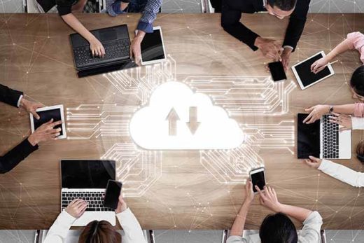 7 people sitting around a large office table using laptops, tablets and smartphone with various white tracing lines coming from them showing a connecting to the cloud uploading & downloading