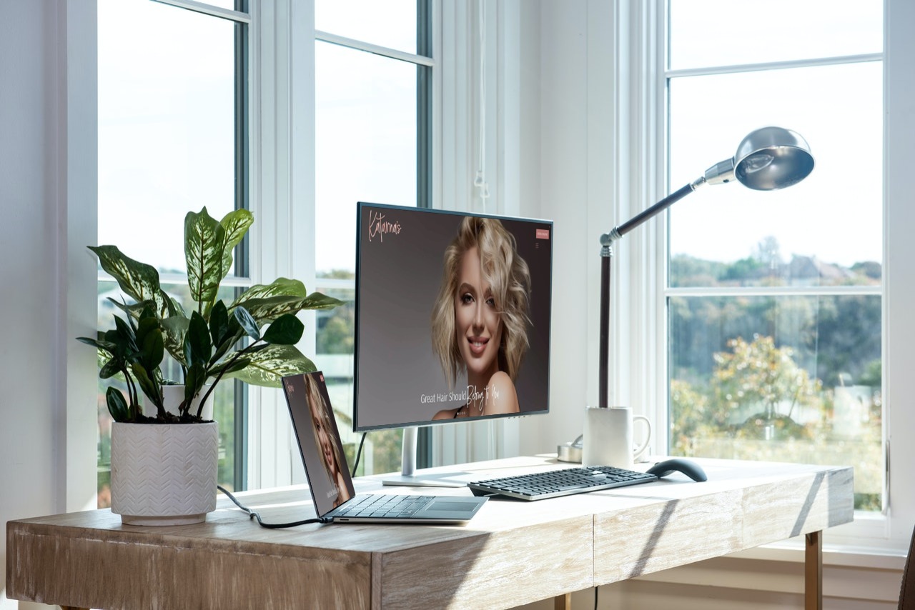 A mockup of the Katarnas Hair Studio website on a desktop and laptop on a desk by windows