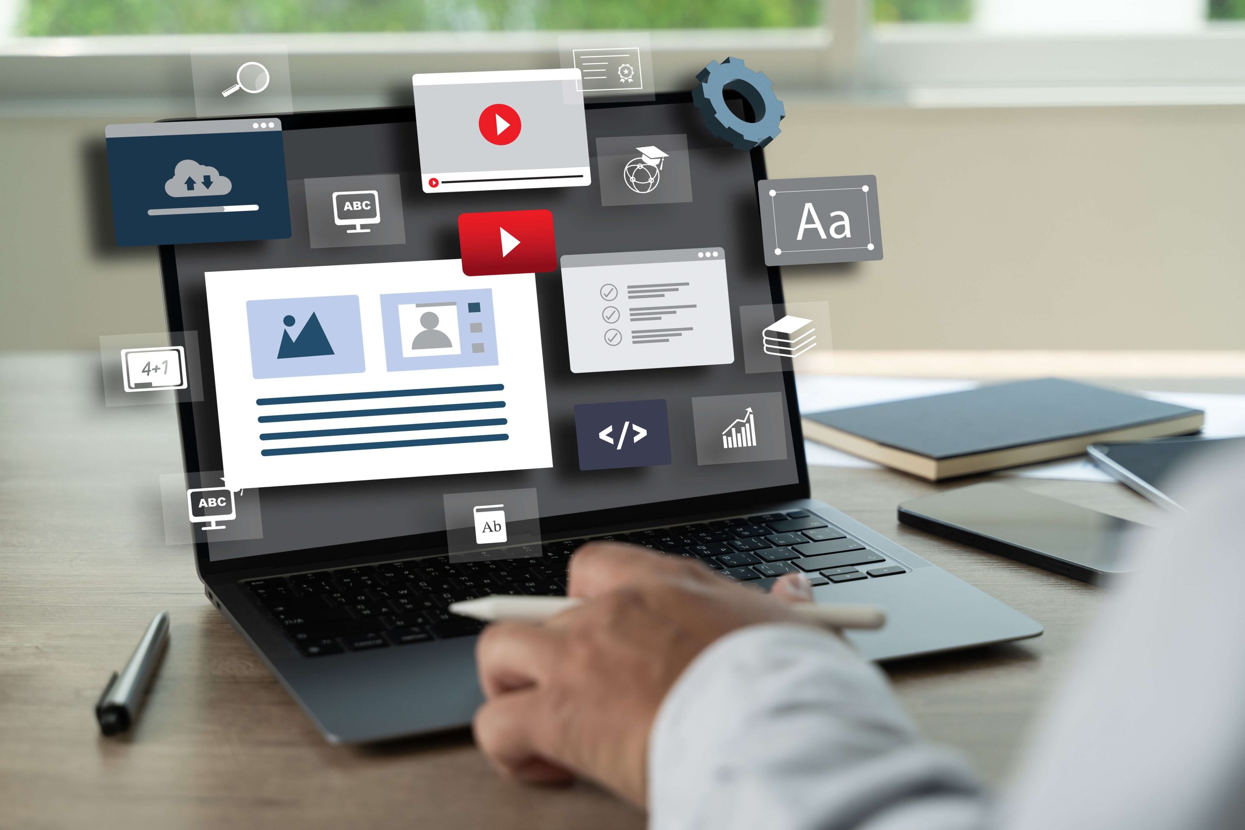 A person using a laptop in a workspace with notebooks and a pen nearby is focused on crafting content for the landing page of a website, displaying digital editing icons for images, text, and video.